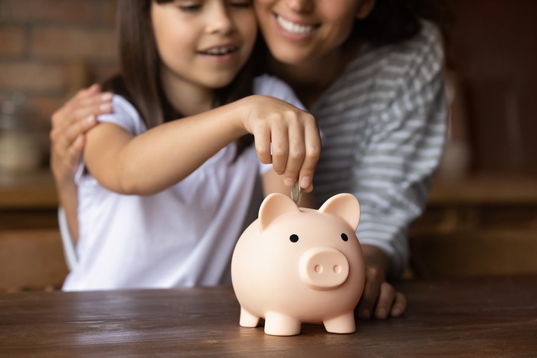 Child putting money into a piggy bank with her mom in the back
