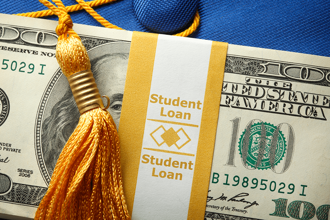 A hundred dollar bill wrapped with paper that says student loans is on top of a graduation cap
