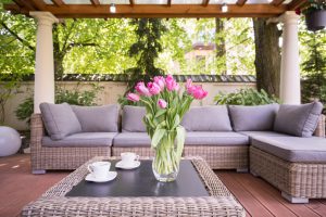 Outdoor living space featuring two white coffee cups and a vase of pink tulips sitting on top a wicker coffee table and surrounded by an L-shaped wicker couch with grey cushions.