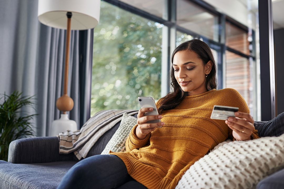 Young woman on her couch on her smartphone while holding a credit card
