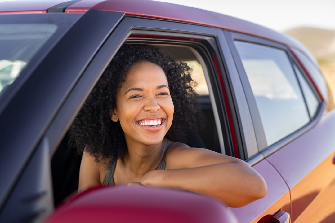 A young woman sits in the driver's seat of a red car, smiling, with her elbow propped on the window.