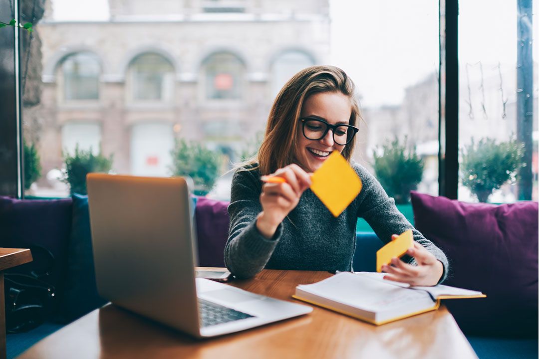 A young woman sitting in a cafe with her laptop and book and booking a stack of sticky-notes.